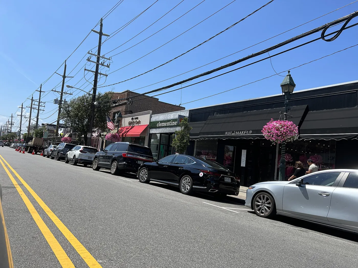 Storefronts along Central Avenue in Cedarhurst on a clear summer morning, with hanging flower baskets and cars parked at the curb.